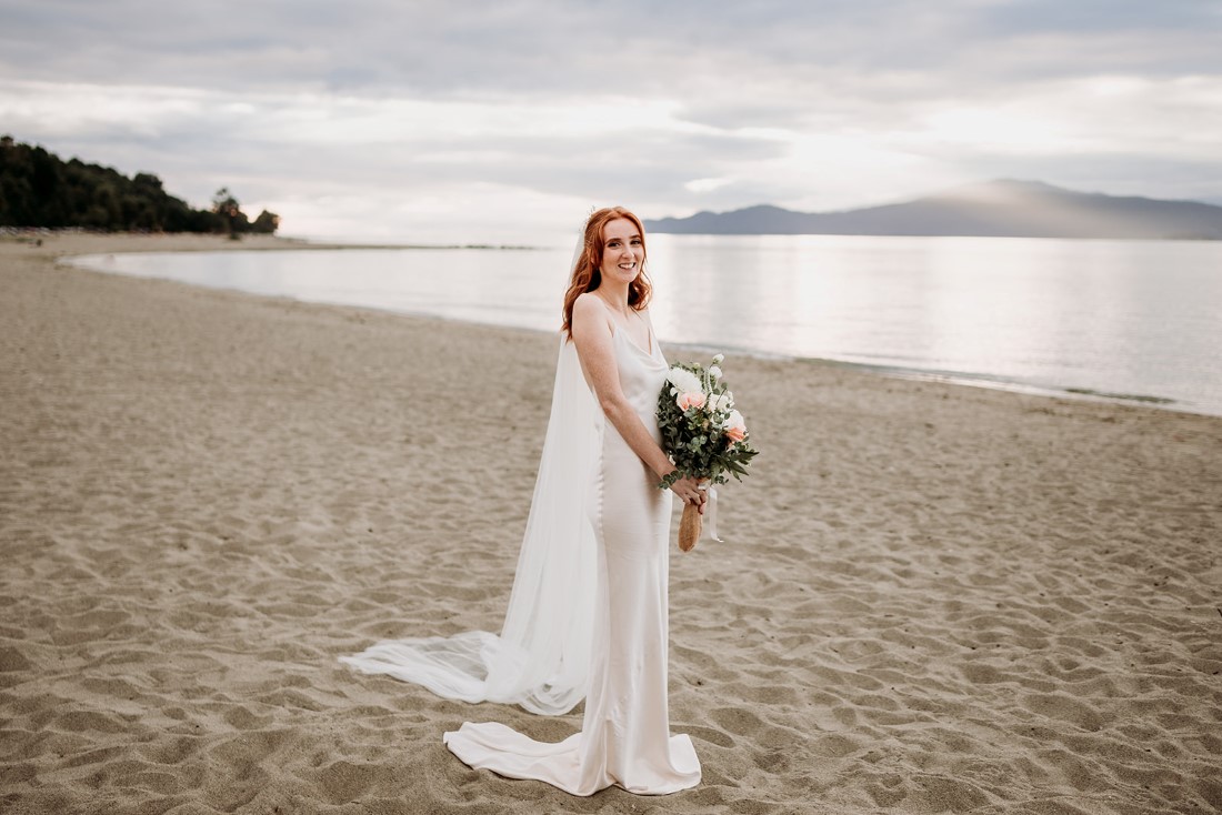 Bride holding bouquet and wearing cathedral veil stands on Vancouver beach