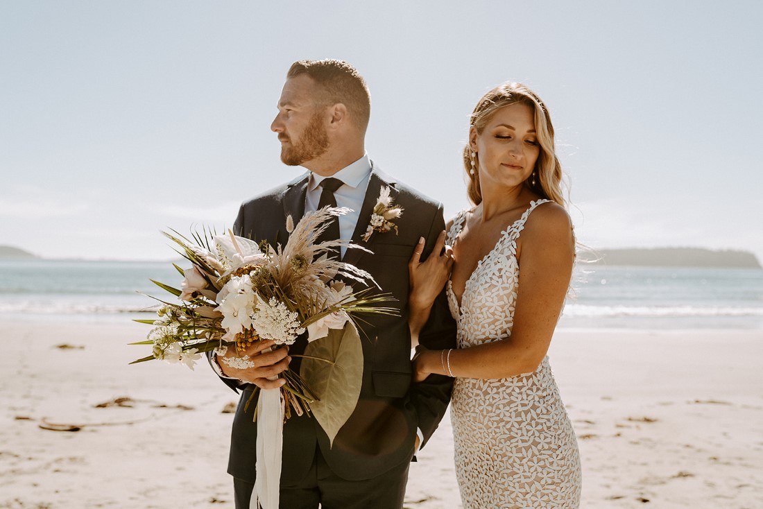 Groom holds Crab Apple Floral bouquet as bride holds on to his arm on Tofino beach