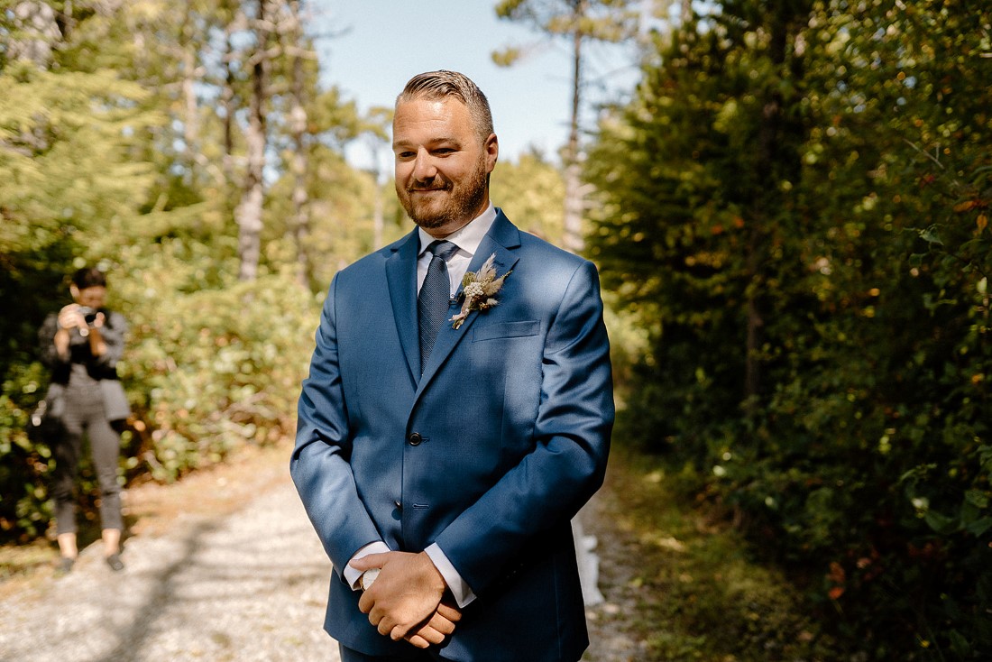 Groom in blue suit smiles at camera on path in Tofino