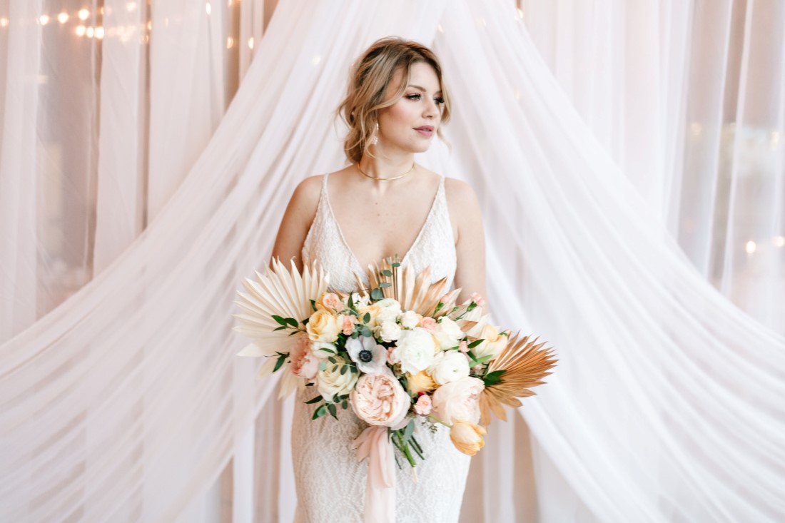 Unbridled Courtney bride stands in front of white drapery holding pastel bouquet