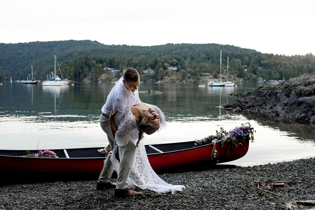 Groom dips his bride in front of a red canoe 