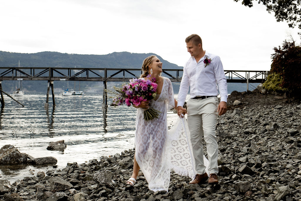 Newlyweds hold hands and walk along the stone beach on Vancouver Island